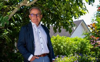 Een man van middelbare leeftijd staat onder een boom met een hand in zijn zak. Hij draagt een wit overhemd en een blauw colbert.