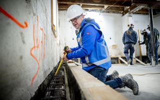 De fundering van een huis wordt onderzocht door een man met een witte helm, een blauwe werkjas en werkschoenen aan.