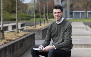Een jongeman met donker haar en een kort baardje zit buiten op een picknicktafel met een boek in zijn hand.