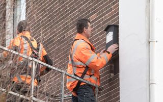 Twee mannen in oranje hesjes staan op een steiger. Die staat tegen een spouwmuur, waaraan een van de mannen een vleermuiskastje bevestigt.