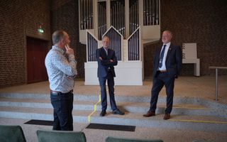 Drie mannen, de twee rechtse in donker pak, met achter hen een orgel met een witte kas.
