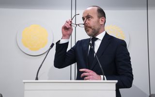 man met bril in zijn hand in een donker pak staat achter een katheder. 