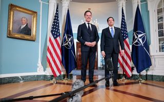 Twee mannen in pak poseren tussen Amerikaanse en NAVO-vlaggen.