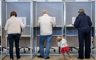 Drie mensen staan in een stemhokje. Een klein meisje gluurt om het hoekje.