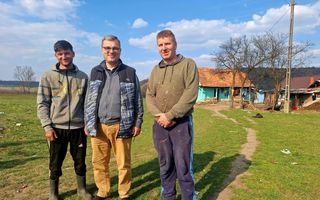 Drie mannen staan naast elkaar op een stuk grasland, met op de achtergrond een huis.