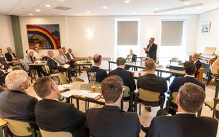 Mannen in donkere pakken luisteren in zaaltje naar spreker, voorin, achter een katheder.