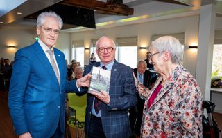 Een man in een blauw pak overhandigt een boek aan de schrijver, terwijl diens vrouw de hand op de rug van haar man legt.