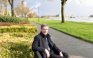 Een man in zwart pak, zittend op een bankje bij een rivier.