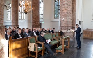 Een man in een donker pak achter een microfoon. Hij staat voor een groep predikanten in een historisch kerkgebouw.