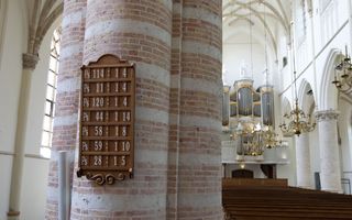 Een houten bord hangt aan een pilaar in een grote kerk. Op het bord staan de psalmen aangegeven die gezongen zullen worden. Verder zijn er bruine banken, witte pilaren en een groot pijporgel te zien.