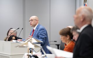 Kale man in lichtblauw pak en rode stropdas achter een spreekgestoelte