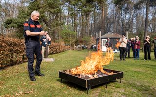 Brandweerman met bril staat naast een vierkante bak met droge bladeren waarin een vuur brandt.