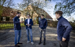 Vier mannen in voornamelijk blauwe kleding staan op het erf van een boerderij met een bloeiende boom ervoor. 
