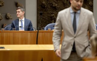 Op de voorgrond man in licht pak, op de achtergrond zit een man in een blauw kostuum.