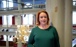 Een vrouw met roodachtig haar, bril in de hand, staand tegen een pilaar van een kerk.