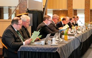 Lange vergadertafel, met daarachter negen mannen, gekleed in donkere pakken.