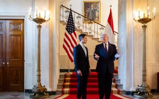 Man met blond haar gebaart naar man met zwart haar; allebei in donker pak op rode loper voor trap met de vlag van Amerika en van Nederland op de achtergrond