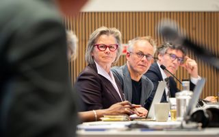 Een vrouw en twee mannen zitten achter aan tafel met een microfoon en kijken schuin opzij,