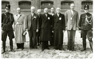 Zwart-wit foto van een groep mannen op een rij voor een gebouw. Ze worden geflankeerd door twee militairen in uniform en hebben allemaal een nummer op de borst gespeld.