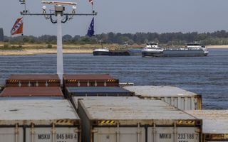 Vanaf een schip dat geladen is met containers kijk je over de rivier naar twee elkaar passerende binnenvaartschepen op de Rijn.