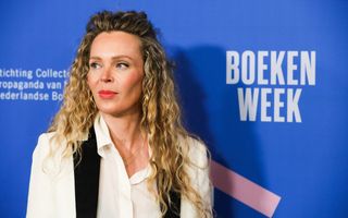 Foto van een vrouw met blonde krullen en een wit blousje aan; ze staat voor een blauwe wand waarop ”Boekenweek” staat.
