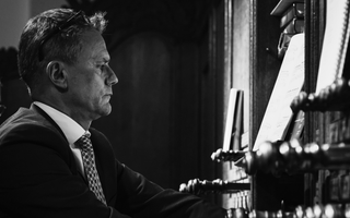 Een man in pak speelt orgel. Links en rechts van hem zijn de registerknoppen van het kerkorgel te zien.