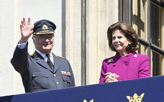 Een man in een uniform zwaait vanachter een blauw kleed waarop kroontjes zijn afgebeeld.