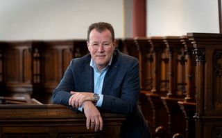 Man met ronde bril en blauw colbert zit op een glanzende houten kerkbank. 