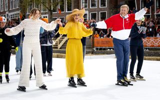 Prinses Ariane met witte trui en broek, koningin M&aacute;xima met gele jurk en hoed en koning Willem-Alexander met blauwe broek en wit-rode trui schaatsen over kunstijs. Op de achtergrond zijn andere schaatsers, publiek en huizen te zien.