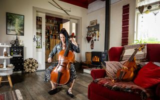 Vrouw met bruin haar zit in een fleurige woonkamer en speelt op een bruin snaarinstrument, dat een barokcello heet. Rechts ligt een ander snaarinstrument op de bank.