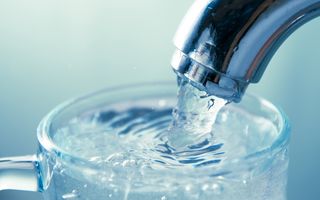 Water stroomt in een glas, uit een kraan waarvan de uitloop rechtsboven te zien is. 
