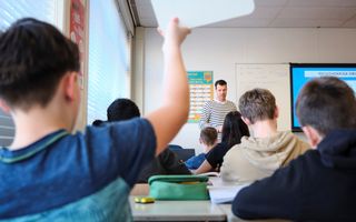 Een leerling steekt zijn vinger op. Voor de klas staat een leraar.