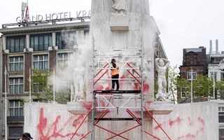 Mannen in oranje hesjes zijn met een hogedrukspuit in de weer voor een groot stenen monument waar rode verf op is gekalkt.