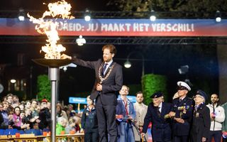 Een man met een ambtsketen om steekt een brandende fakkel in een schaal. Ernaast staan twee veteranen, tussen hen in een man met een witte pet.