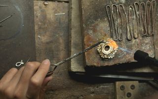 Ruwe stenen, ijzerdraad. Een hand is bezig met pincet en brandertje om een fijn, gouden sieraard te maken. 