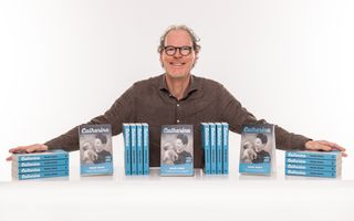 Foto van een man met krullend haar en bril. Voor hem staan blauwe boeken 