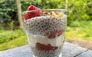 Een glaasje op een houten tafel waarin chiapudding, rode rabarberjam en witte yoghurt in laagjes te zien zijn.