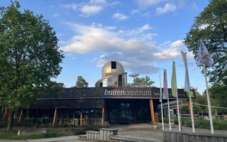 Laag houten gebouw met daarbovenop een toren met een zilverkleurige koepel. Links en rechts staan bomen met groen blad. In de lucht zijn enkele grijs-witte wolken te zien.