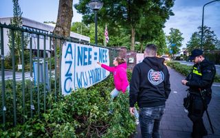 Een vrouw met blond haar en roze jas steekt haar hand uit naar een wit spandoek op een hek met als tekst: "azc, nee=nee, wij houden vol".