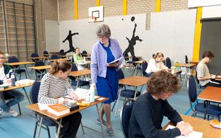 Een vrouw in een blauwe jurk loopt met een stapel papieren op de arm langs tafeltjes waaraan jongren zitten.