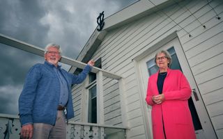 Ouder echtpaar voor een wit houten huis met het symbool voor geloof, hoop en liefde erop. De man heeft een blauwe trui aan, de vrouw een roze jurk.