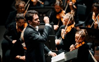 Een dirigent in een zwart pak staat met opgeheven rechterarm tussen de musici van een orkest.
