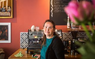 Vrouw met donker haar en een blauwgroen truitje onder een zwart vest staat voor een koffiemachine.