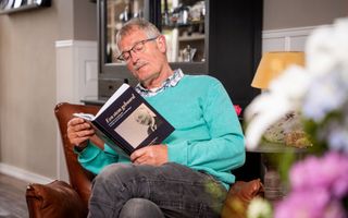Een oudere man met een azuurblauwe trui in een bruine stoel; hij zit een boek te lezen.