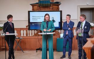 Vrouw staat in groen broekpak vlak voor een preekstoel tussen mannen in pak