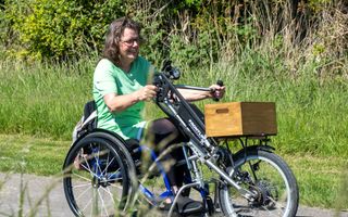 Vrouw in een groen shirt rijdt op een handbike met een houten kist voorop langs frisgroen gras.
