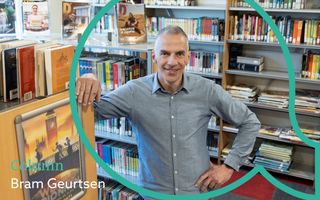 man van middelbare leeftijd met lichtblauw overhemd staat in een bibliotheek, tussen kasten vol boeken