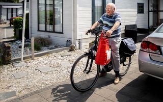 Breed lachende oude heer zit op de fiets op zijn oprit. Aan het stuur van zijn fiets hangt een rode boodschappentas.