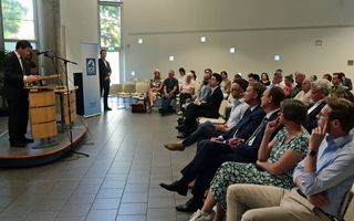 Links een man in pak achter een katheder. Rechts mensen op stoelen die luisterend naar hem kijken.