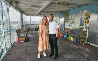 Een vrouw met een jurk en een man met een baard en een wit overhemd aan boord van een schip. Op de achtergrond twee houten banken en een beschilderde wand.